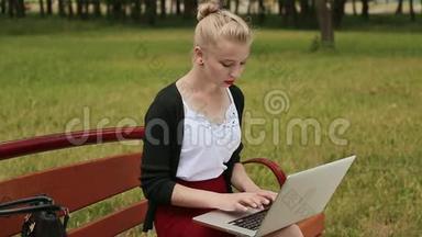 美丽的年轻女孩，金发放在公园的长凳上，在她的笔记本电脑上工作。 女孩用笔记本电脑打字。 侧视。